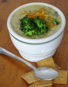 broccoli cheddar soup in w hite bowl with green lines and a spoon and wheat thin crackers. 