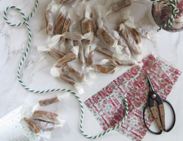 flatly on white marble with a christmas tree green and white twine cellophane bags sissors and homemade caramels wrapped in waxed paper aa-2