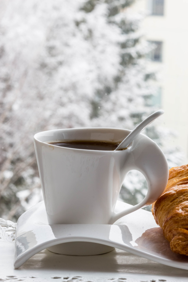 cup of chocolate and a croissant on a white plate in front of a snowy window