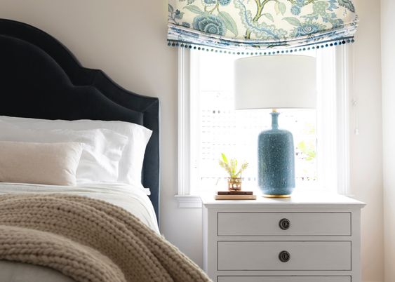 Bedroom with a blue upholstered headboard, white side tables and blue pattern roman shades. 