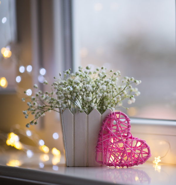 woven pink heart by a window with fairy lights and a white pot of flowers.