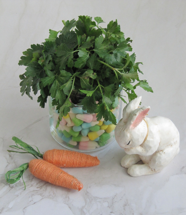 Easter rabbit decoration with a clear glass vase filled with pastel easter candy and herbs and two small carrots sitting on the side. 