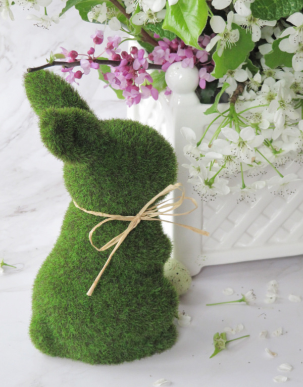 Green moss rabbit sitting next to a white lattice vase with flowers.