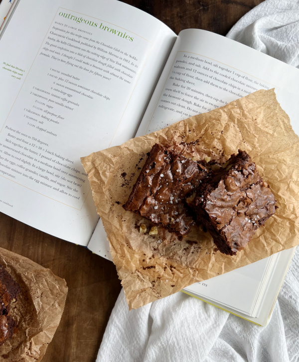 Weekend Meanderings two brownies on a brown piece of parchment sitting on an open cookbook with the recipe for outrageous brownies.