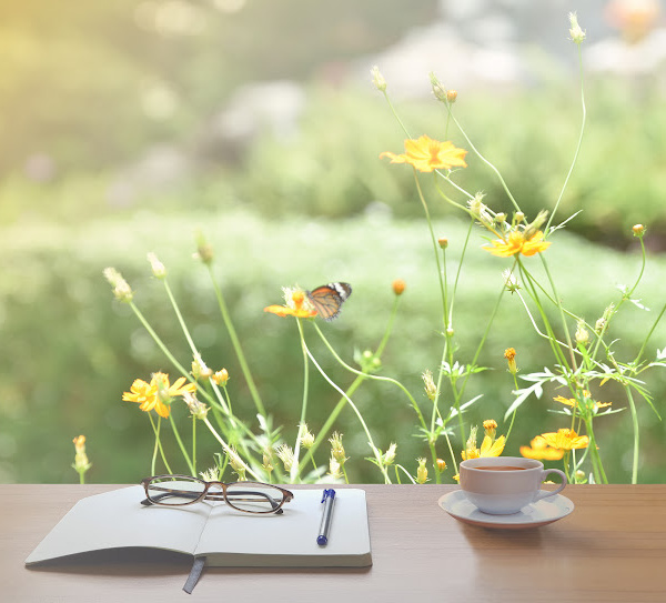 cup of tea in a white cup sitting next to a notebook with a pen and glasses with yellow flowers and a butterfly