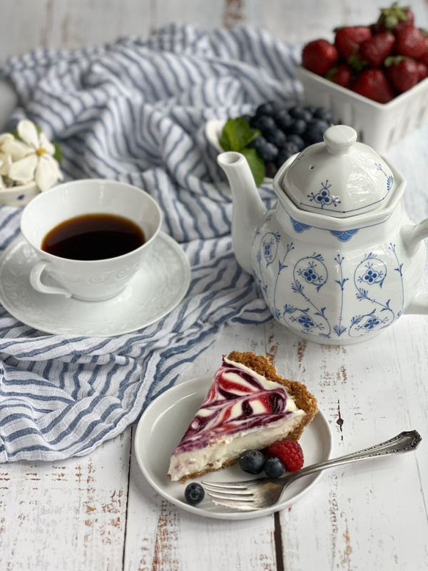 blue and white tea pot, cup of tea, piece of cheesecake and a basket of blueberries and strawberries.