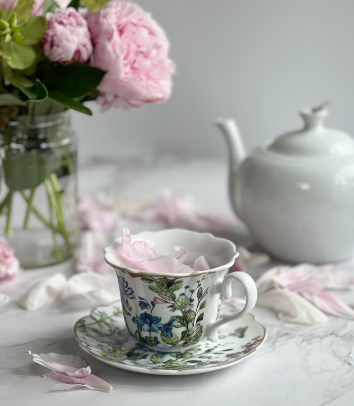 pink peonies in a glass vase with a white teapot and a teacup with pastel flowers filled with peony petals.