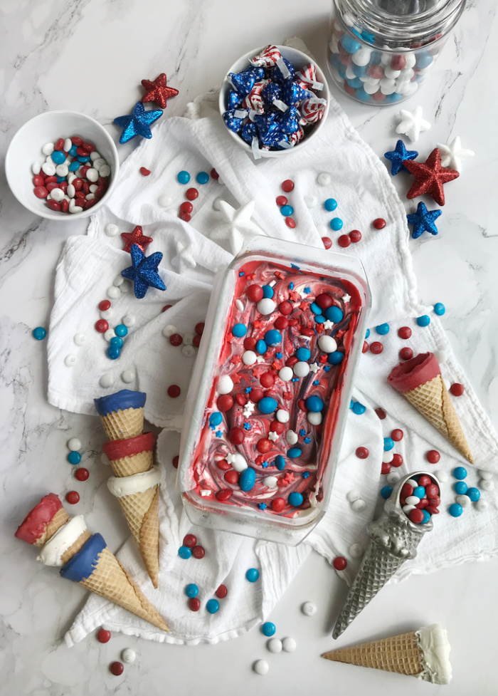 Red White Blue No churn ice cream in a loaf pan with sugar cones dipped in red, white and blue chocolate and bowls of patriotic candy. 