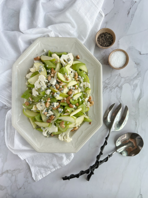 Celery Apple Cauliflower salad on a white ironstone platter with two small wood bowls of salt and pepper and a pair of serving utensils. 