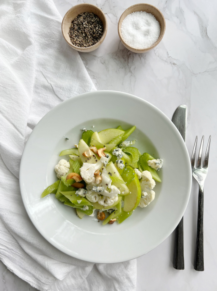 celery salad in a bowl with salt and pepper in two small bowls and a fork and knife.