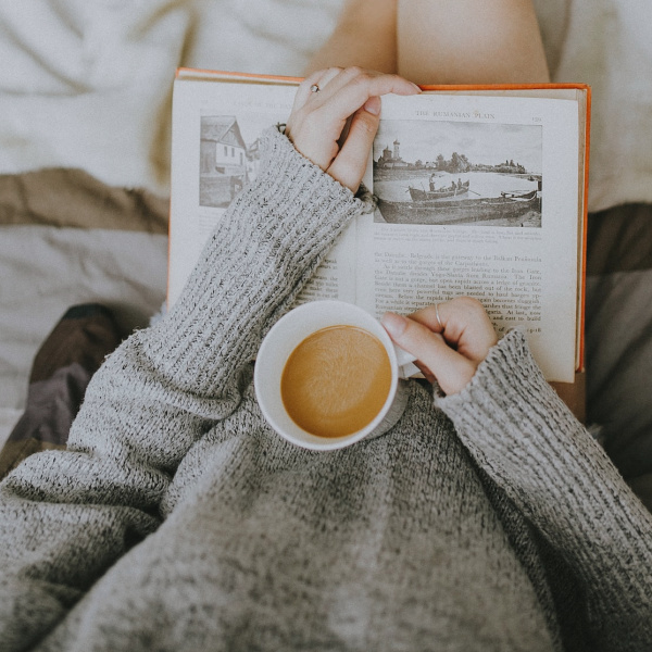 Woman wearing a gray sweater, reading a book and drinking a cup of coffee. 