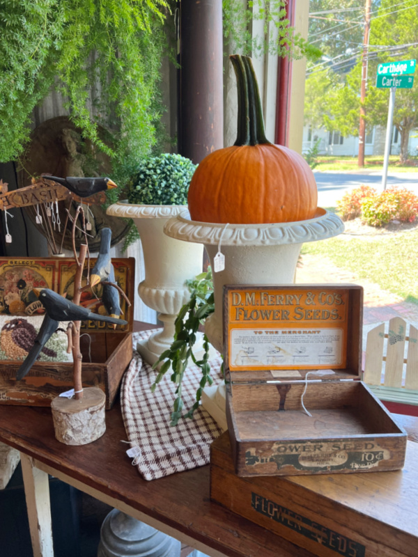 Large orange pumpkin in a white urn on a table filled with antiques.