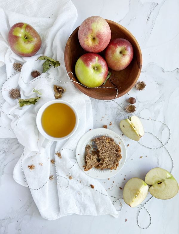 three apples in a bowl with a slice of applesauce bread and a cup of tea.