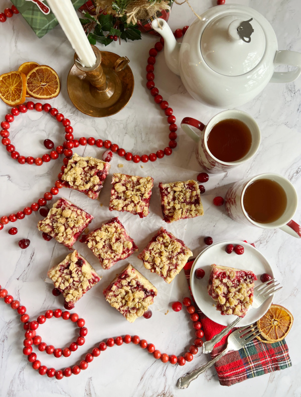 Flatlay of cranberry bars, pot of tea and cup with a mini Christmas tree and dried orange slices.