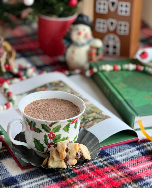 white cup with holly filled with hot chocolate with some christmas books and a gingerbread house