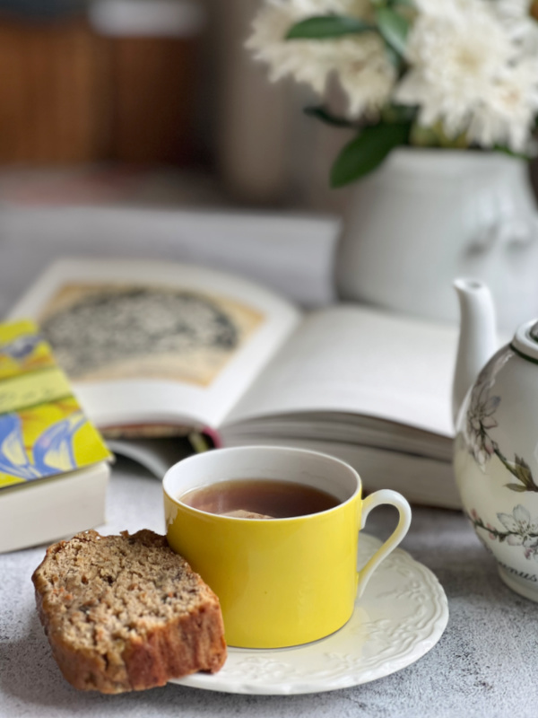 tea in a yellow cup with a slice of banana bread and 2 books with a teapot and a vase of flowers.