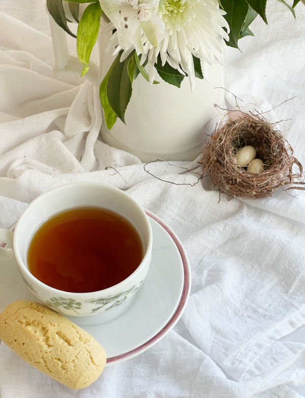 white pitcher of flowers, cup of tea and biscuit and a small bird nest with 2 eggs.