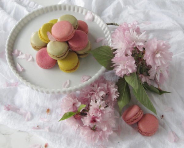 Pastel French Macarons and pink cherry blossoms sitting in on w white cake platter.
