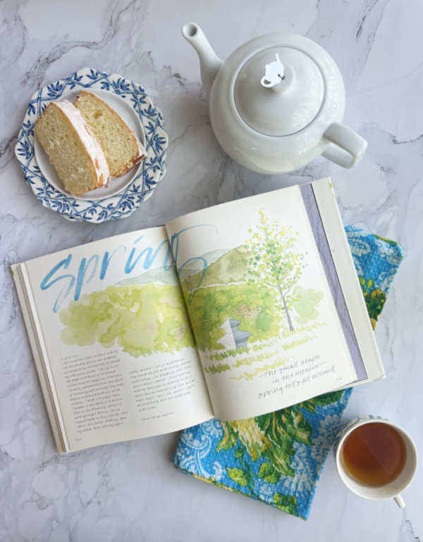 Spring Flatlay with a white tea pot, a cup of tea, a spring book and tw slices of lemon bread on blue and white plates.