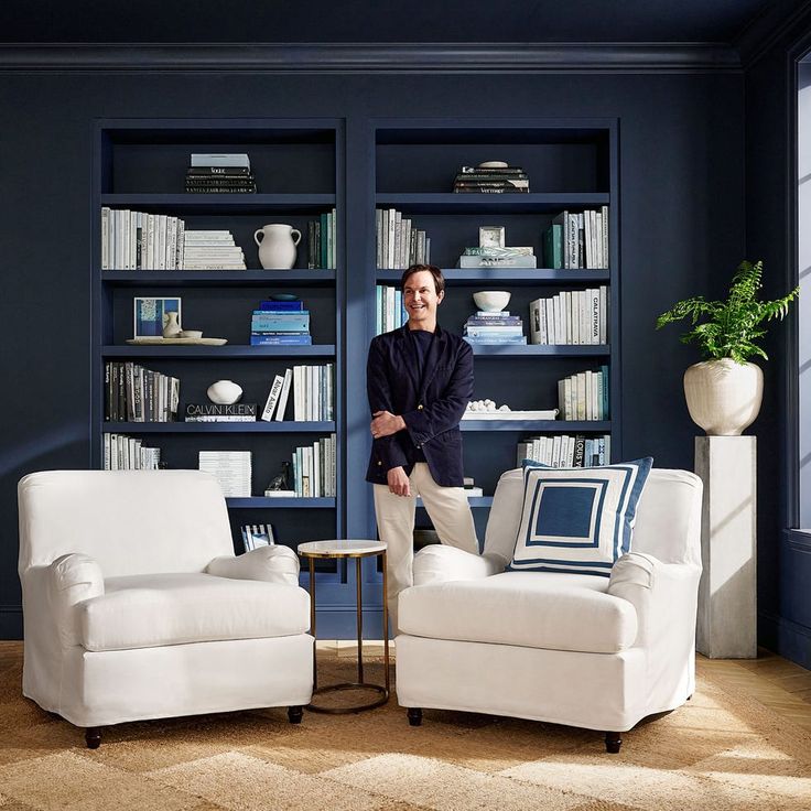 Mark D sikes x Pottery barn blue room with book cases and white chairs and a man in white pants and blue blazer. 