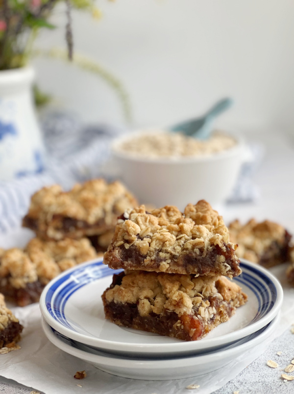 Old Fashioned Date Bars stacked on a plate with a bowl of oats and a blue scoop in the background.