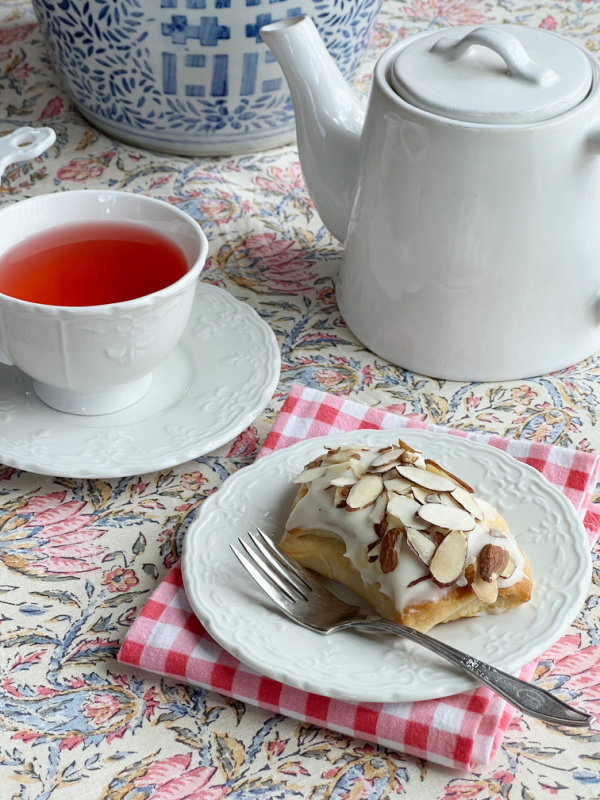 almond bear claw danish pastry on a white plate sitting on a pink and white gingham napkin with a pot of tea and a cup.