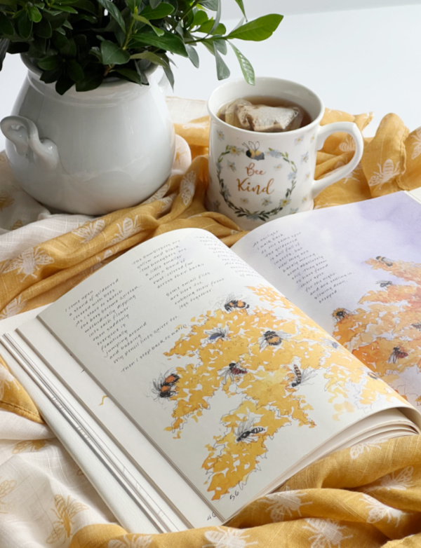 Bee kind flatlay with mug of tea, yellow and white bee scarf and a book with illustrations of bees.
