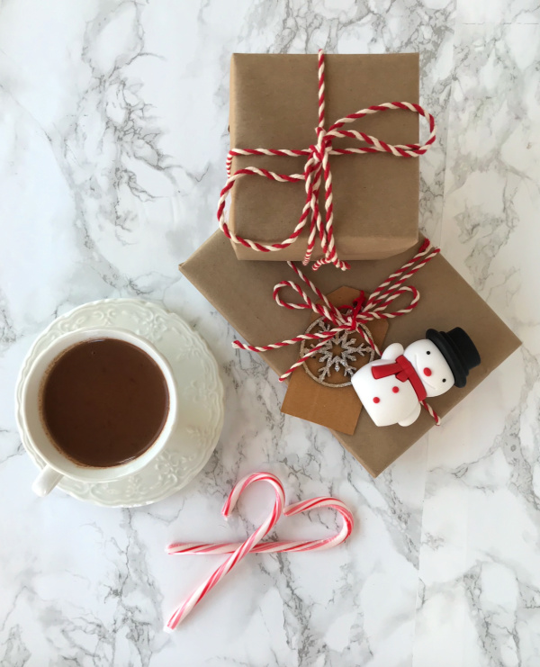 cup of hot chocolate and two brown wrapped packages with red and white twine and a small snowman and candy canes.