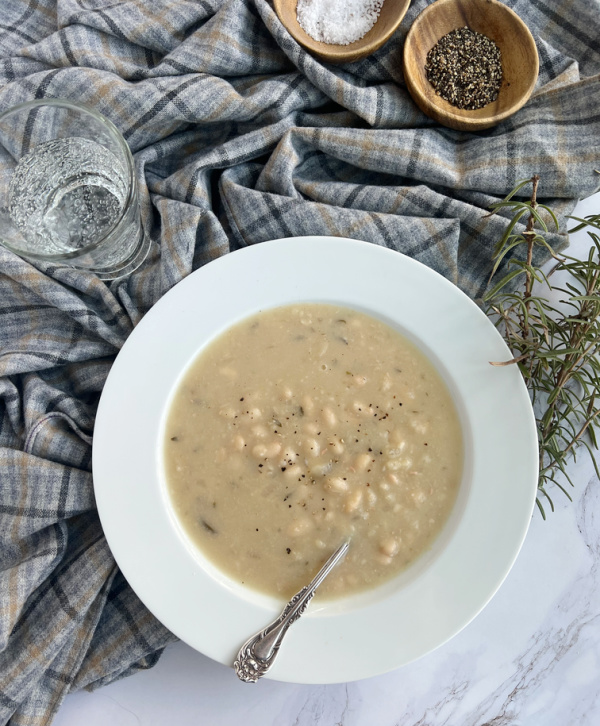 Barefoot Contessa Rosemary white bean soup in a white bowl with a plaid tablecloth, a small wood bowl of salt and pepper and a glass of sparkling water.