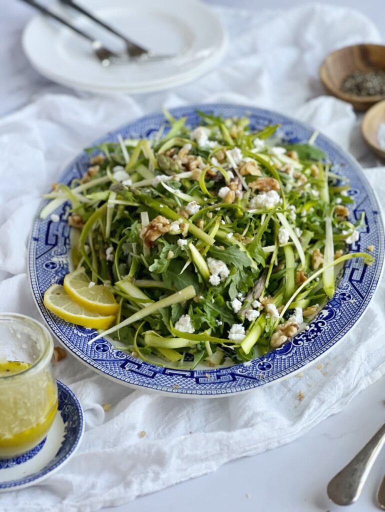 shaved asparagus salad with lemon vinaigrette feta and walnuts on a blue and white serving platter. and a jar of lemon vinaigrette.
